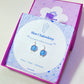 Blue Chalcedony Petite Round Stud Earrings displayed on crystal meaning card inside purple Laganna jewelry box - handcrafted wire-wrapped healing jewelry gift from Sri Lanka
