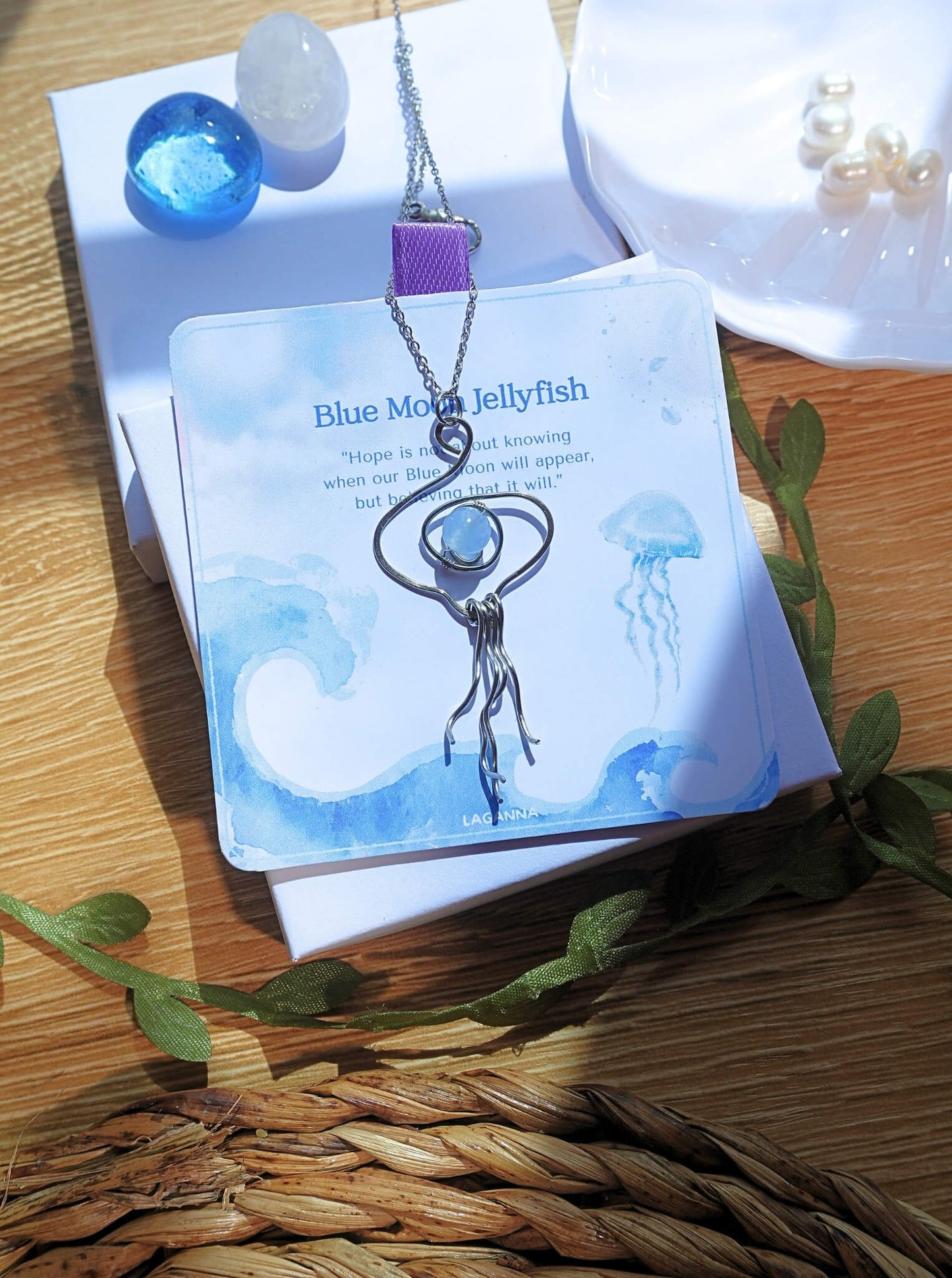 Blue Moon Jellyfish Necklace handmade stainless steel wire jellyfish pendant with Blue Chalcedony on Laganna packaging card angled view wooden surface artisan crafted Sri Lanka healing crystal jewelry tarnish resistant water resistant