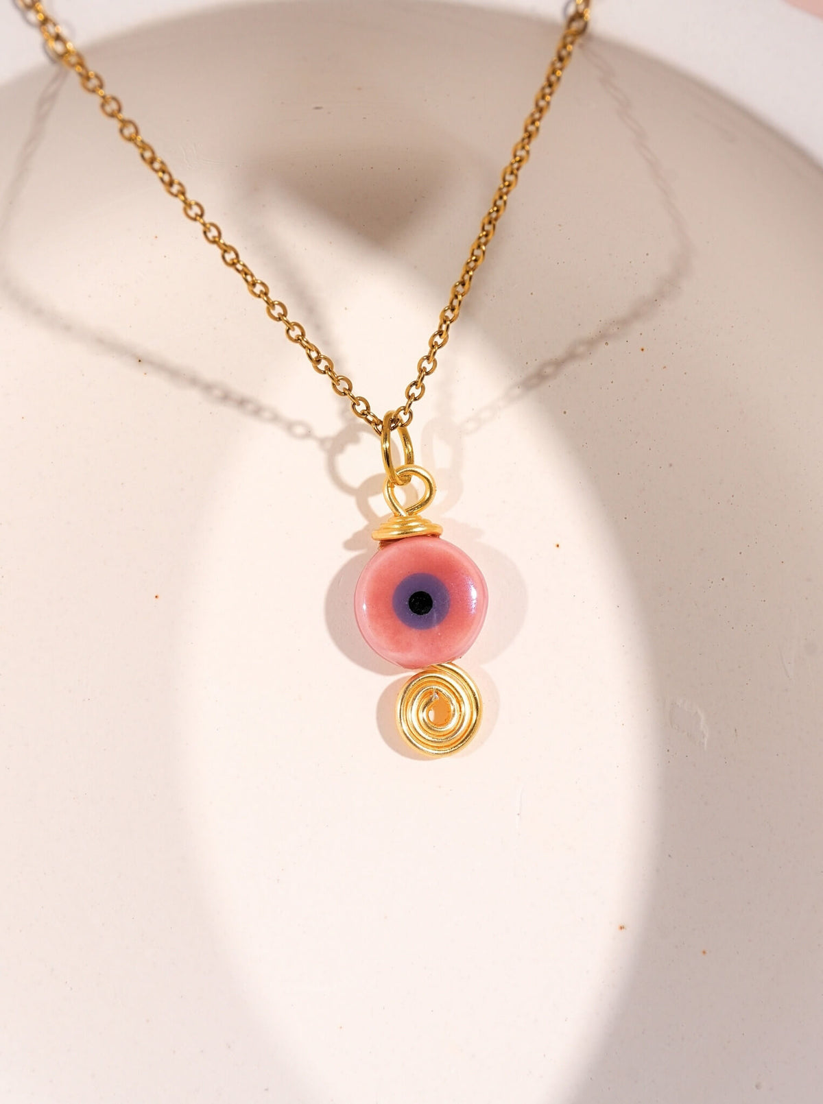 Gold pink evil eye protection necklace with ceramic bead and spiral charm on white display, handmade Sri Lankan spiritual jewelry featuring wire wrapped evil eye pendant as self-love talisman and compassion amulet for women seeking emotional balance and heart healing
