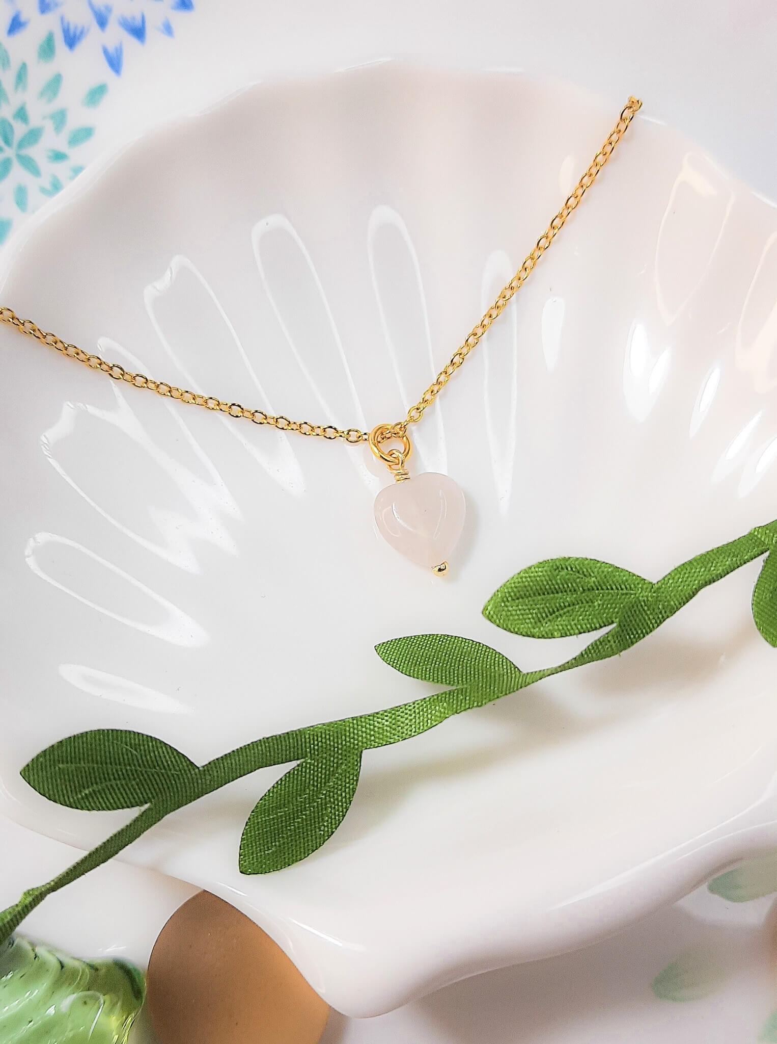 Sweet heart rose quartz necklace on white dish with natural leaves showing handmade gold wire wrapped healing crystal jewelry, Sri Lankan artisan heart chakra pendant featuring heart-shaped pink gemstone perfect as boho love necklace and self-love amulet for nature-loving jewelry enthusiasts