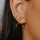 Wire-wrapped bow earrings in gold plated finish worn on ear close-up, handmade artisan jewelry by Laganna Sri Lanka, delicate feminine bow design with graceful wire fall, lightweight stud earrings for everyday wear, available with or without Japanese Miyuki Delica glass beads in powder blue and cream beige, premium handcrafted accessories for women