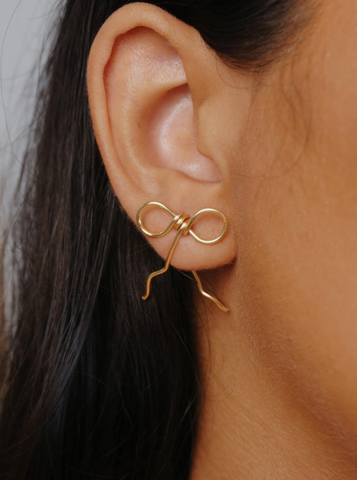 Wire-wrapped bow earrings in gold plated finish worn on ear close-up, handmade artisan jewelry by Laganna Sri Lanka, delicate feminine bow design with graceful wire fall, lightweight stud earrings for everyday wear, available with or without Japanese Miyuki Delica glass beads in powder blue and cream beige, premium handcrafted accessories for women