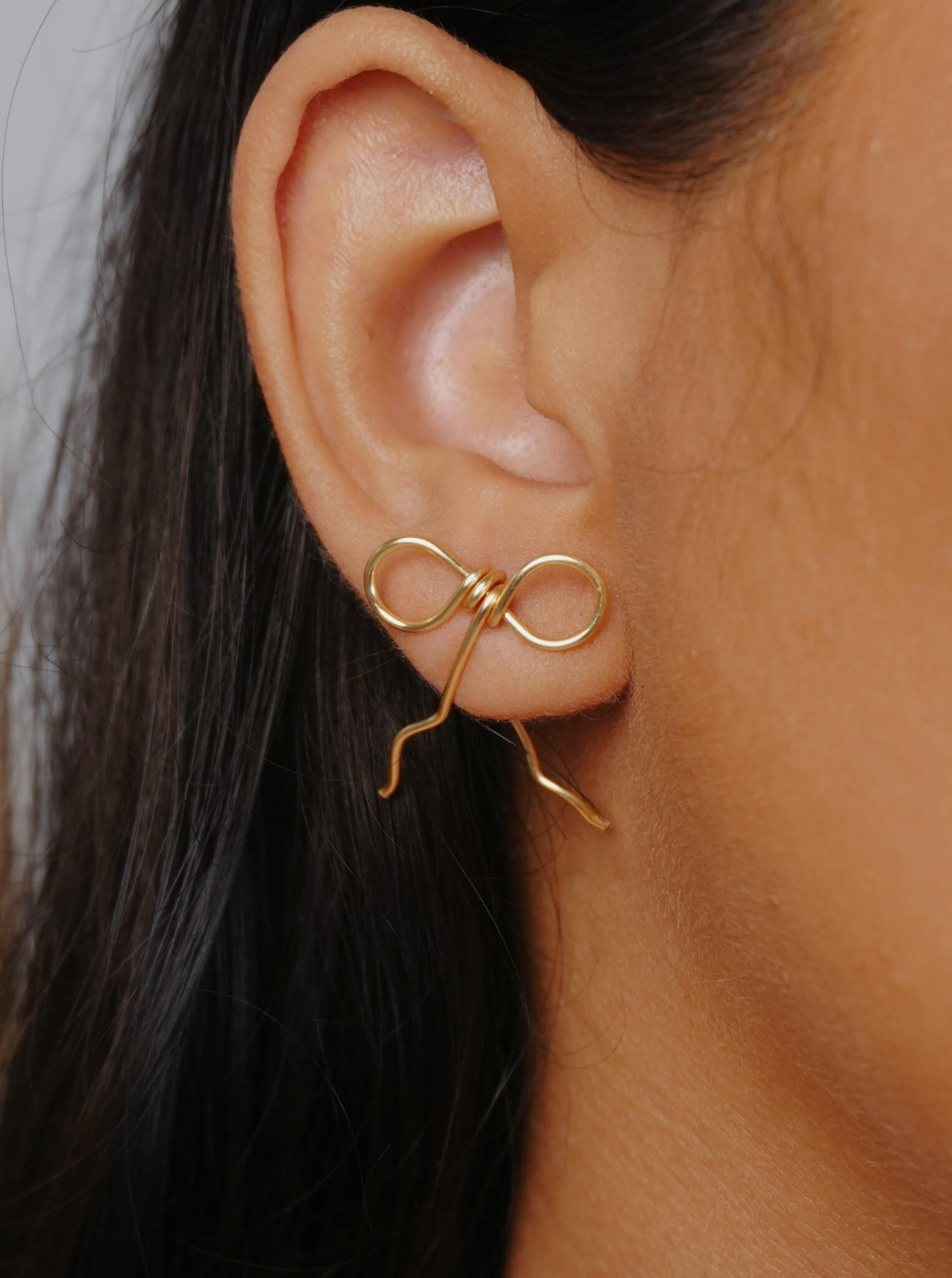 Wire-wrapped bow earrings in gold plated finish worn on ear close-up, handmade artisan jewelry by Laganna Sri Lanka, delicate feminine bow design with graceful wire fall, lightweight stud earrings for everyday wear, available with or without Japanese Miyuki Delica glass beads in powder blue and cream beige, premium handcrafted accessories for women